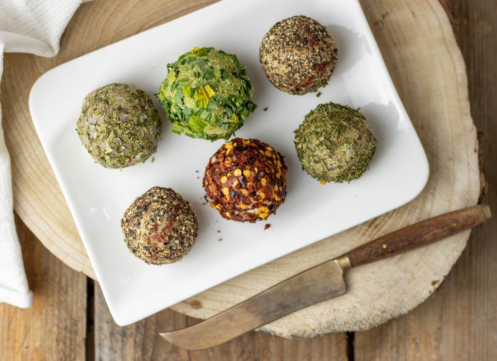 6 vegane, cremige Frischkäsebällchen auf Cashewbasis auf einem länglichen Teller.
