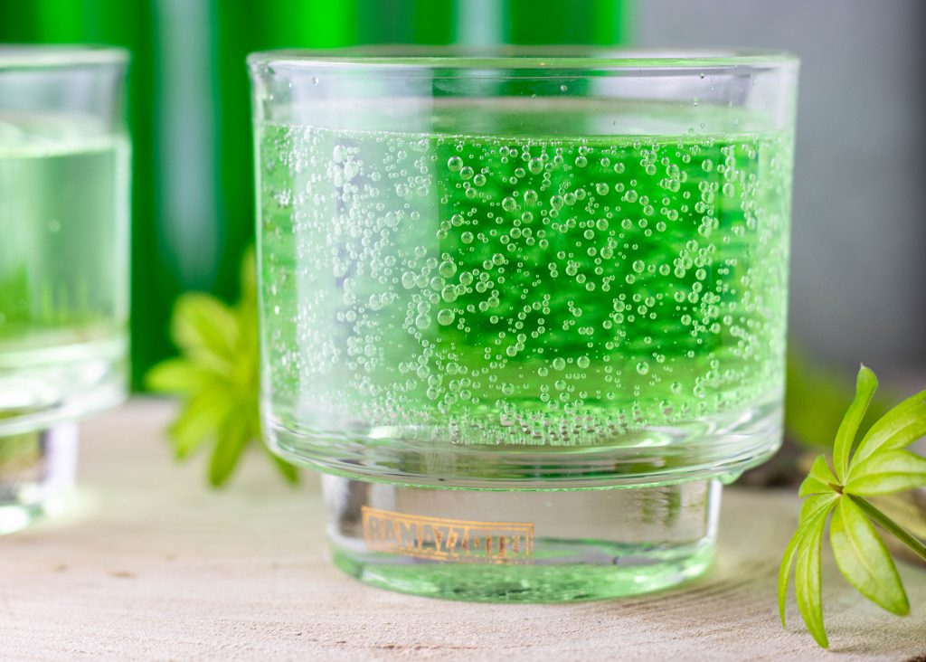 Sprudelige Waldmeisterbrause im Glas, daneben frischer Waldmeister.