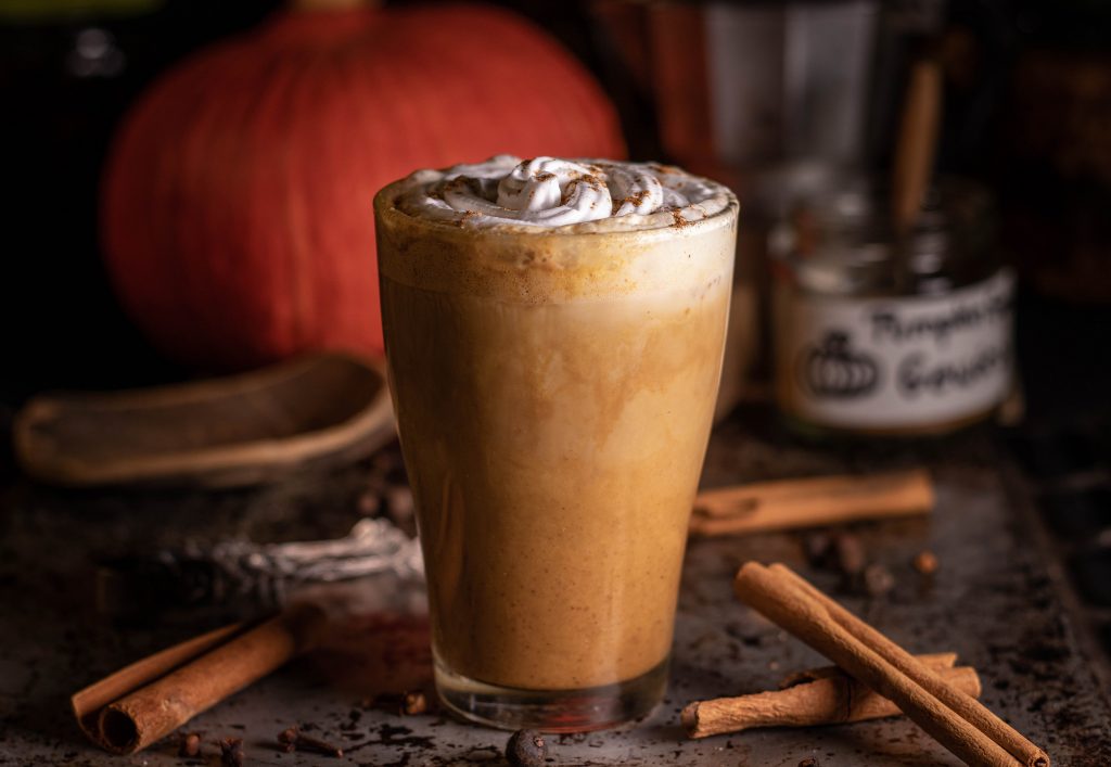 Pumpkin Spice Latte in einem Glas, darum Zimtstangen und ein Kürbis