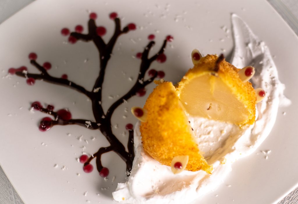 In Cider pochierte Birne. Ausgebacken im knusprigen Kokosmantel! Serviert auf duftendem Tonkaschaum und einem Schokoladen-Rotweincreme-Bäumchen.