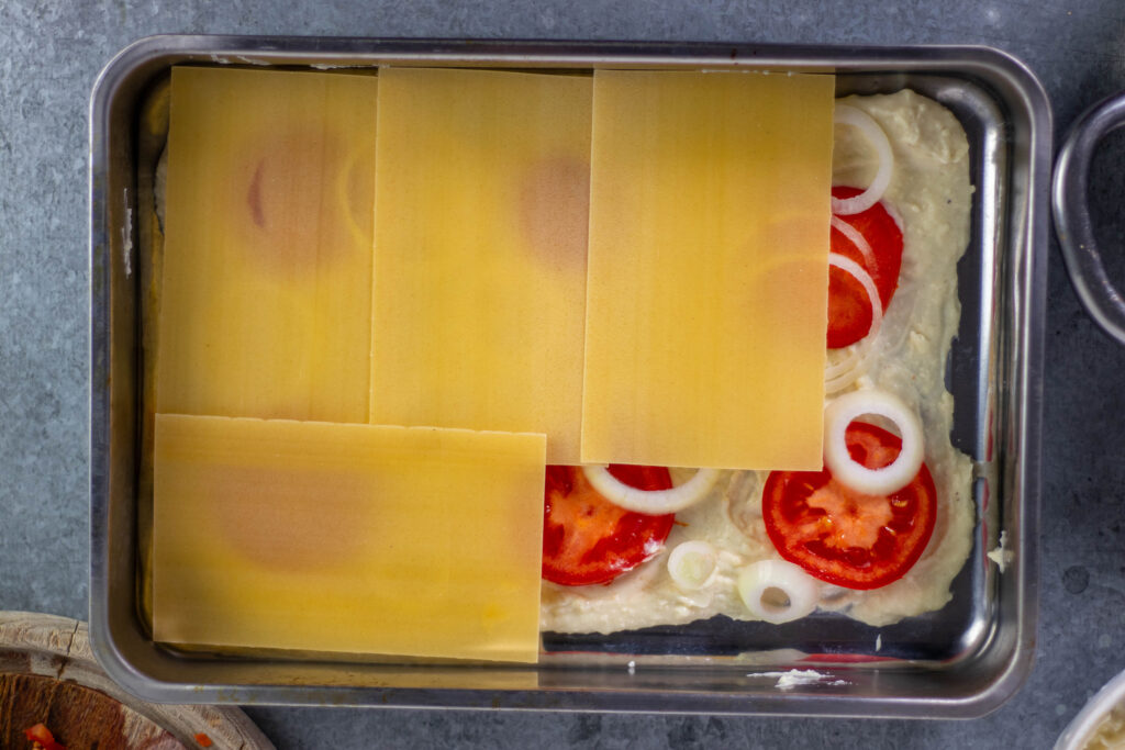 7. Die unterste Schicht der veganen Lasagne besteht aus Bechamelsauce mit Tomatenscheiben und Zwiebelringen. Darauf kommen die ersten Lasagneblätter.