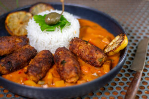 Vegan Ćevapčići with bell pepper sauce, raosted lemons and rice on a black plate