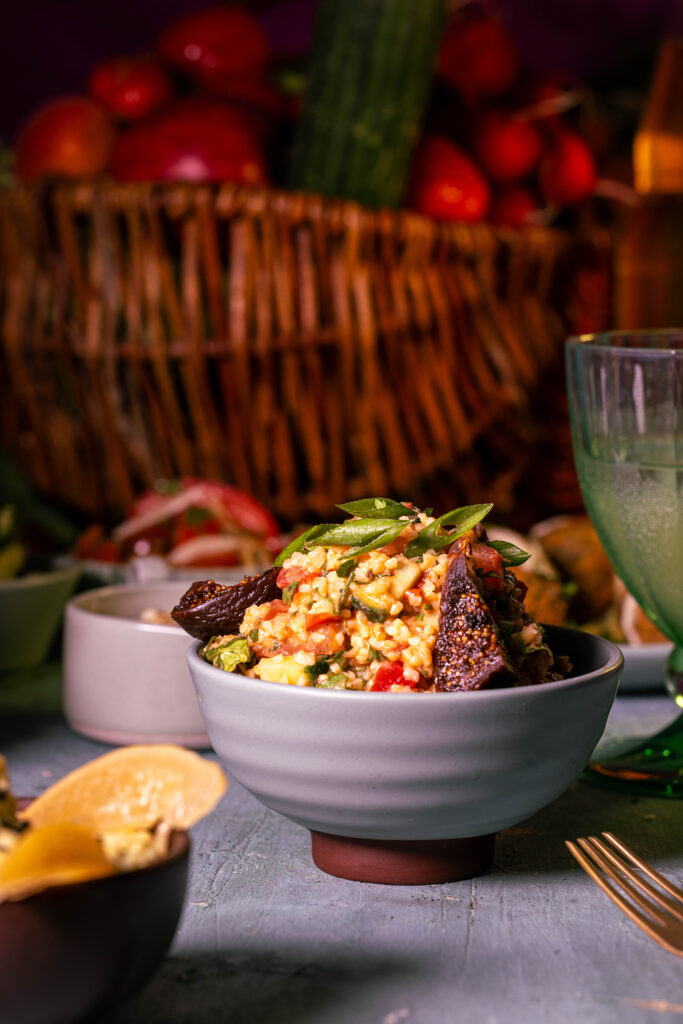 Bulgursalat mit Kefir und getrockneten Feigen auf einem Tisch mit einem großen Korb voller Obst und Gemüse, sowie kleinen Beilagen.