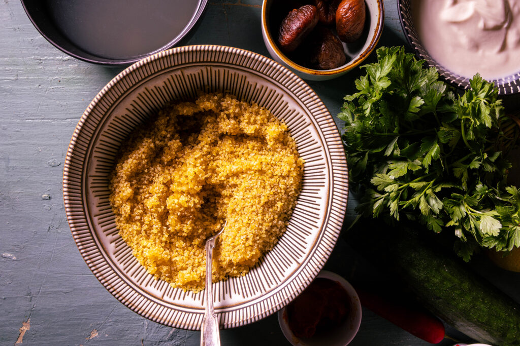 Eine Schale mit Bulgur und einem Löffel darin. Daneben weitere Zutaten.