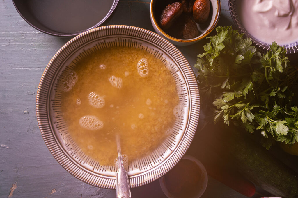 Eine Schale mit Bulgur und dampfendem, heißen Wasser. Mit einem Löffel darin. Daneben weitere Zutaten.