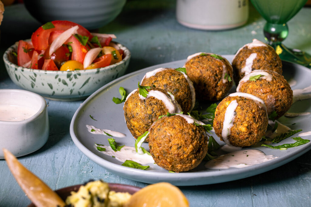 Falafel mit Tahini-Soße auf einem Teller. Daneben Tomatensalat und eine Schale Tahini-Soße.