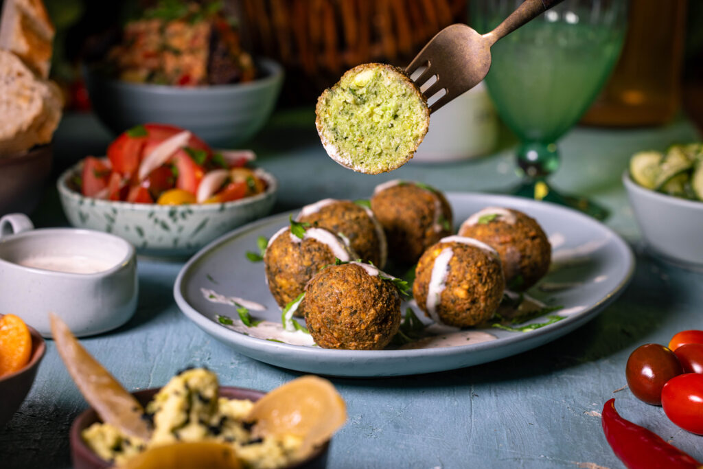 Falafel auf einem Teller. Einer ist halbiert und aufgespießt auf einer Gabel. Drumrum verschiedene Beilagen.