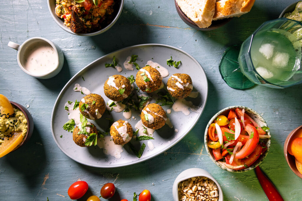 Falafel von oben auf einem Teller. Mit Tahini-Soße besperenkelt. Drumrum verschiedene Beilagen.