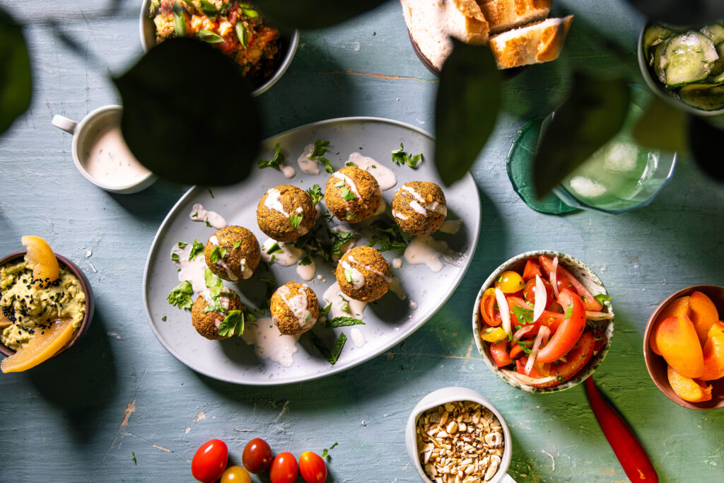 Falafel von oben auf einem Teller. Mit Tahini-Soße besperenkelt. Drumrum verschiedene Beilagen.