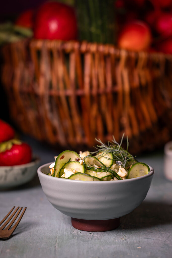 Gurkensalat mit Kokoskefirdressing in einer Schale, dahiner ein Korb mit Obst.
