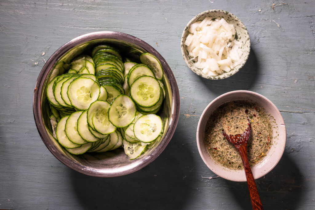 Eine Schale mit geriebener Gurke, eine mit fein geschnittener Zwiebel und eine mit Dressing