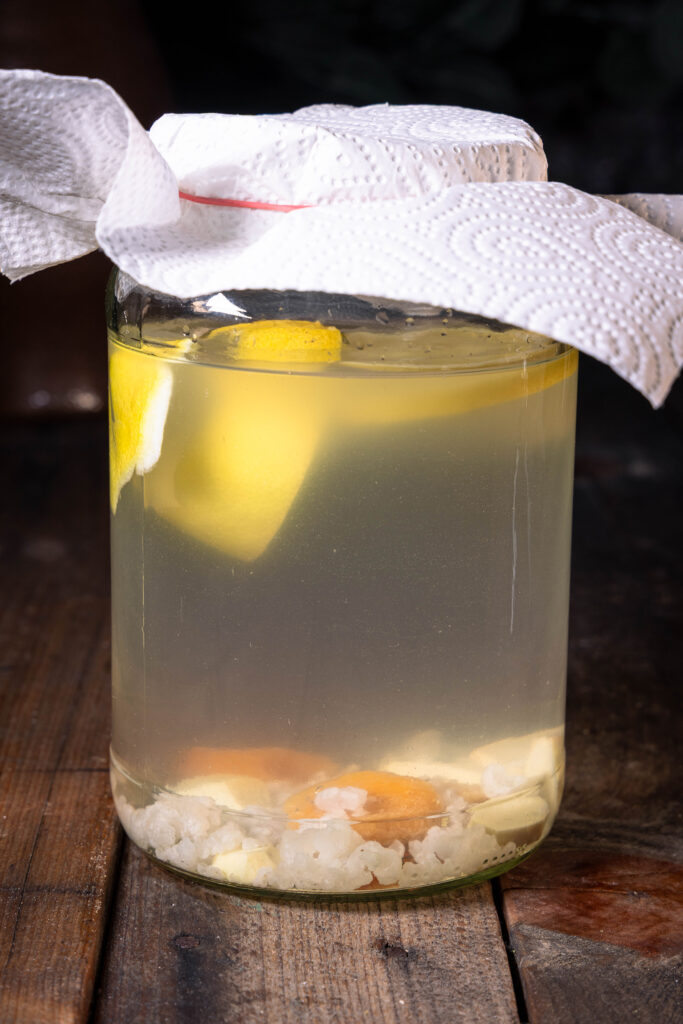 Wasserkefir fermentiert in einem großen Glasgefäß, das mit einem Küchenpapier abgedeckt ist.