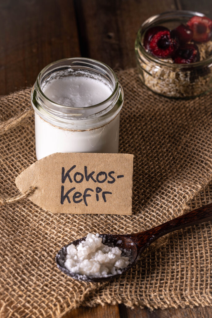 Kokoskefir in einem Glas, deneben eine Schale Müslig mit frischen Kirschen und eine Löffel mit Wasserkefirkristallen.