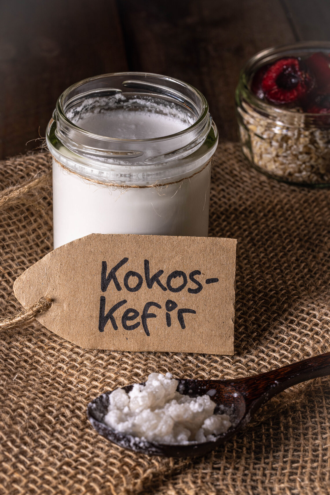 Kokoskefir in einem Glas, deneben eine Schale Müsli mit frischen Kirschen und eine Löffel mit Wasserkefirkristallen.