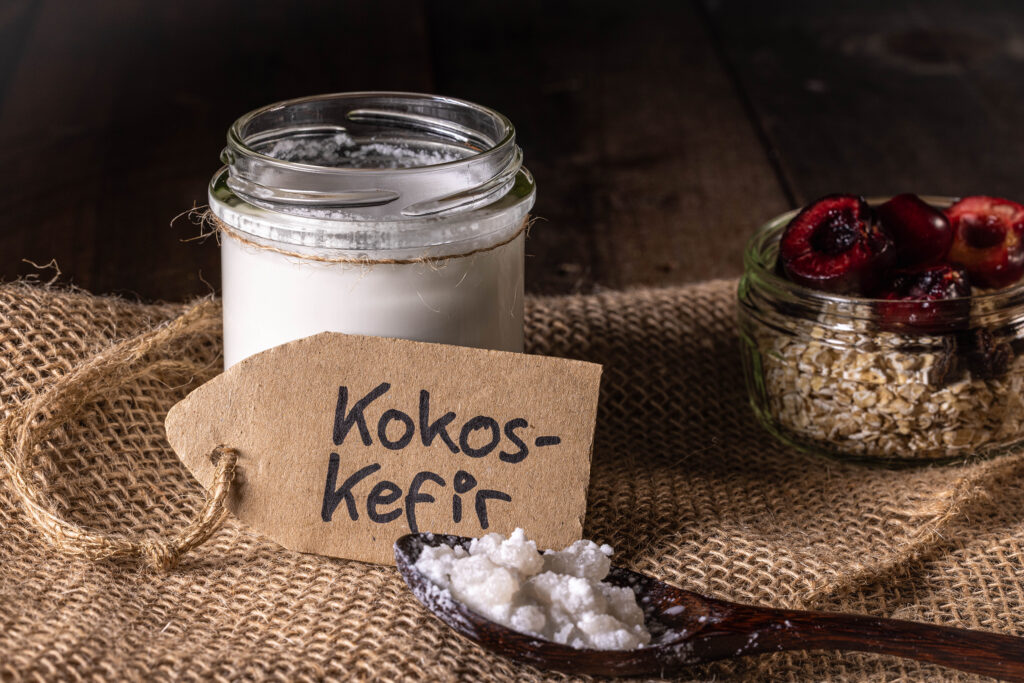 Kokoskefir in einem Glas, deneben eine Schale Müslig mit frischen Kirschen und eine Löffel mit Wasserkefirkristallen.