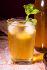 Kombucha-Grundrezept - eine Glas mit Kombucha, einem Stengel Melisse und Eiswürfeln. Dahinter eine Flasche mit Kombucha.