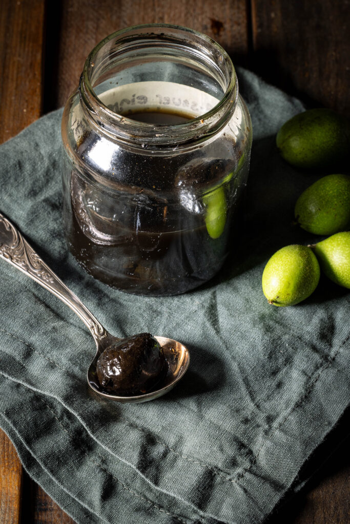 Süß-sauer eingelegte Schwarze Nüsse – im Glas und auf einem Löffel. Daneben frische unreife Walnüsse vom Baum.
