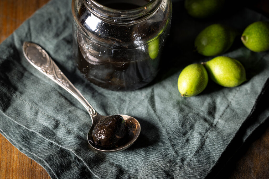 Süß-sauer eingelegte Schwarze Nüsse – im Glas und auf einem Löffel. Daneben frische unreife Walnüsse vom Baum.