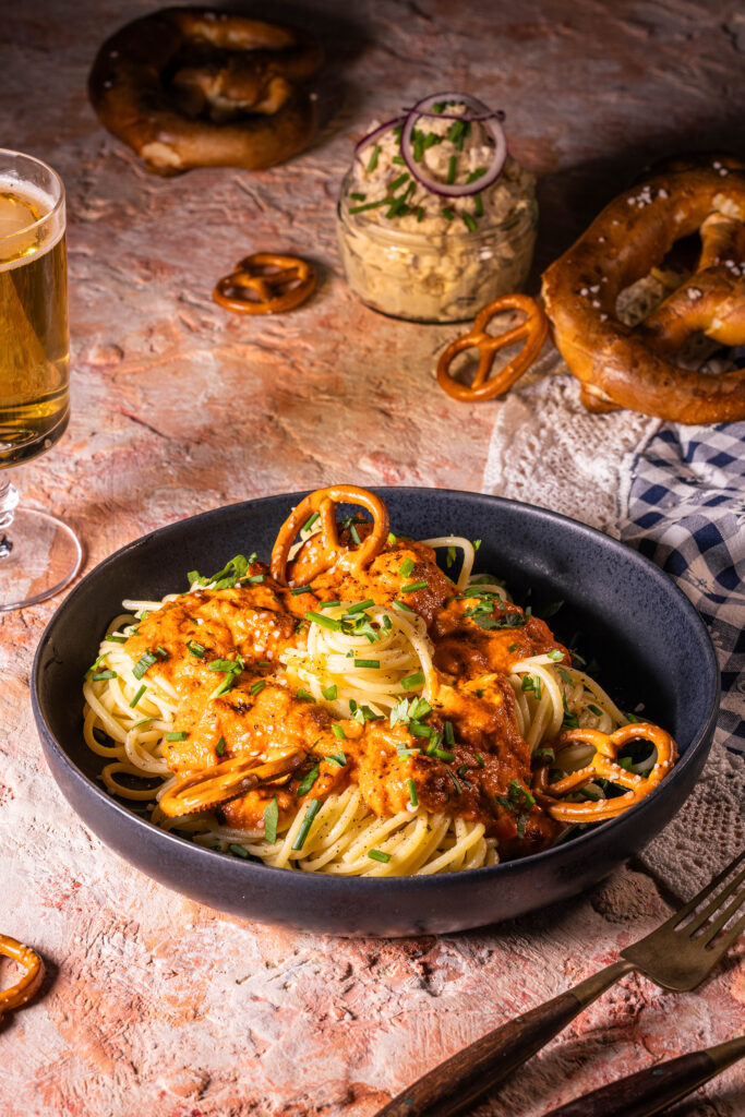 Spaghetti al Camembert – Pasta mit Obazda-Sauce. Auf einem Teller angerichtet. Dahinter Brezeln, eine Schale Obazda und Bier.