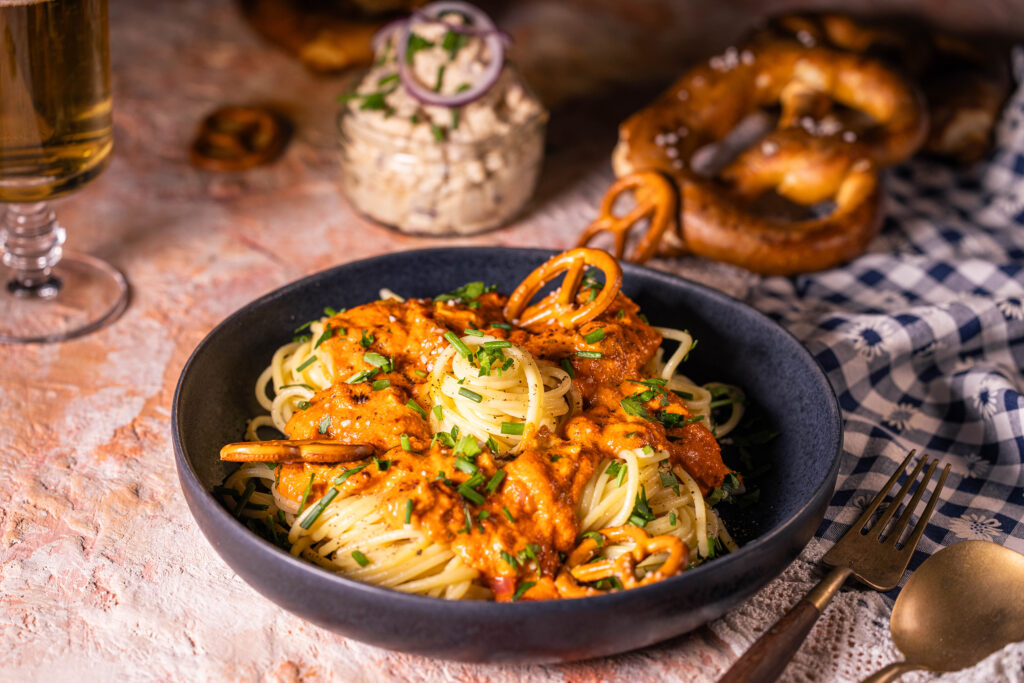 Spaghetti alla Camembert – Pasta mit Obazda-Sauce - vegan. Angerichtet auf einem Teller. Dahinter veganer Obazda, Brezeln und Bier.