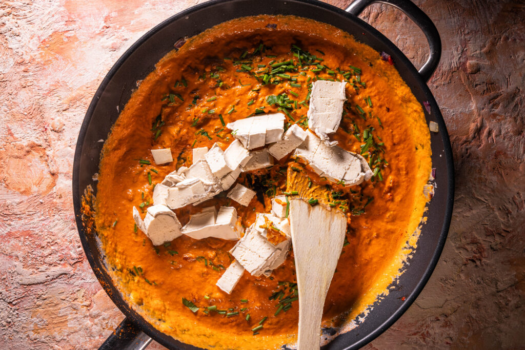 Veganer Camembert, Petersilie und Schnittlauch werden in eine rote Sauce in einer Pfanne gegeben.