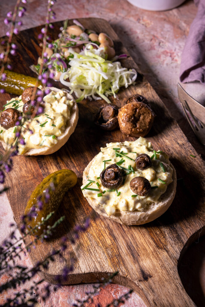 Zwei Brötchenhälften mit veganem Eiersalat auf einem Holzbrett. Außerdem weitere Beilagen.