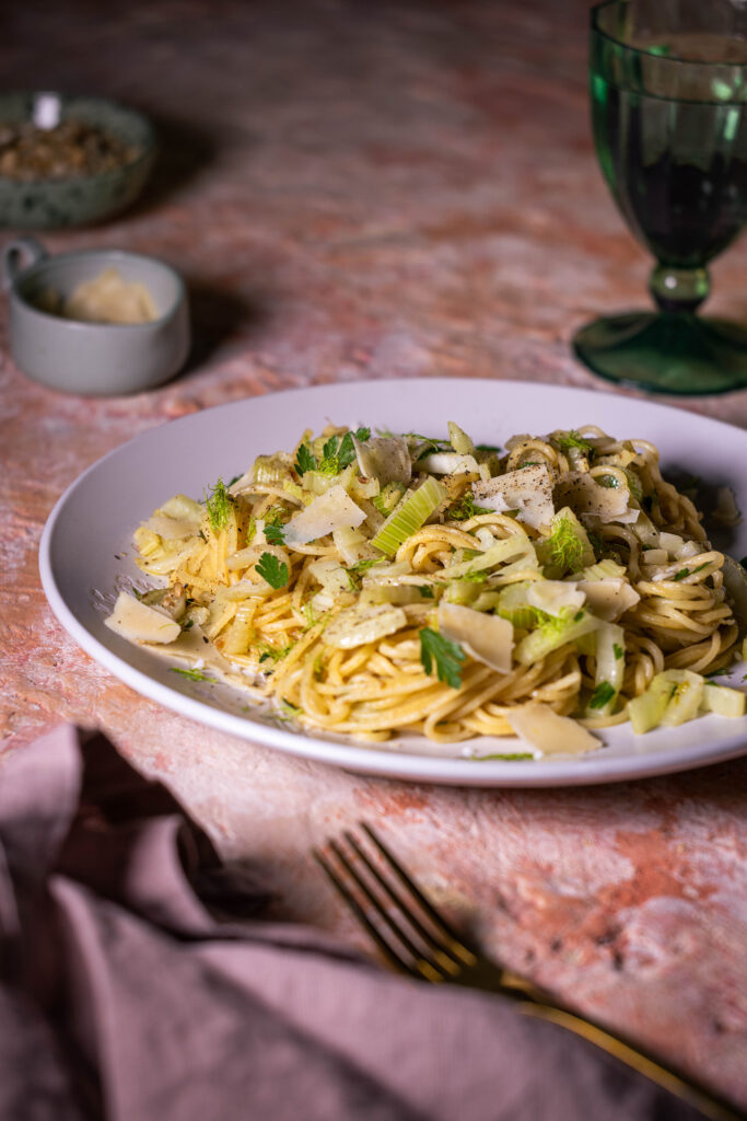 Fenchel Spaghetti, mit Knoblauch und Zitrone, darauf Parmesan gestreut. Auf einem hellen Teller, davor Gabel und Serviette, dahinter ein Glas und kleine Schalen.