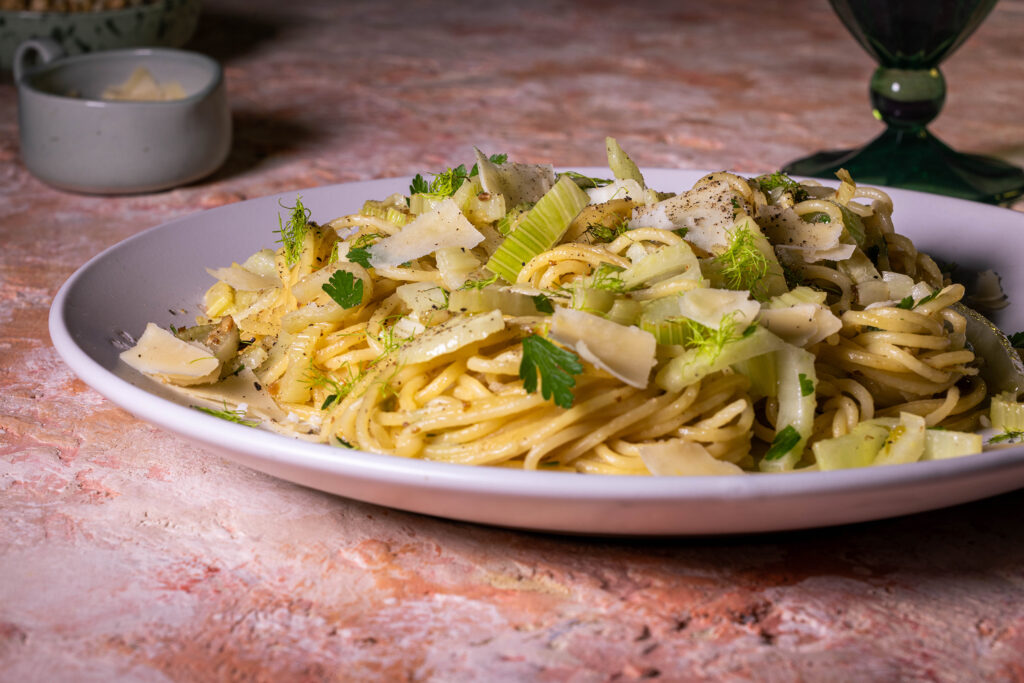 Fenchel Spaghetti, mit Knoblauch und Zitrone, darauf Parmesan gestreut. Auf einem hellen Teller, dahinter ein Glas und kleine Schalen.