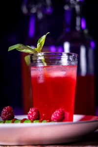 Himbeer-Kardamom-Kombucha in einem Glas mit Eiswürfeln. Dahinter Kombucha in Flaschen. Davor frische Himbeeren.