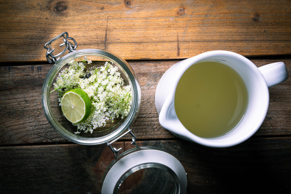 Holunderblüten, Limette und Holundersirup in einem verschließbaren Glas. Daneben eine Karaffe mit Kefir.