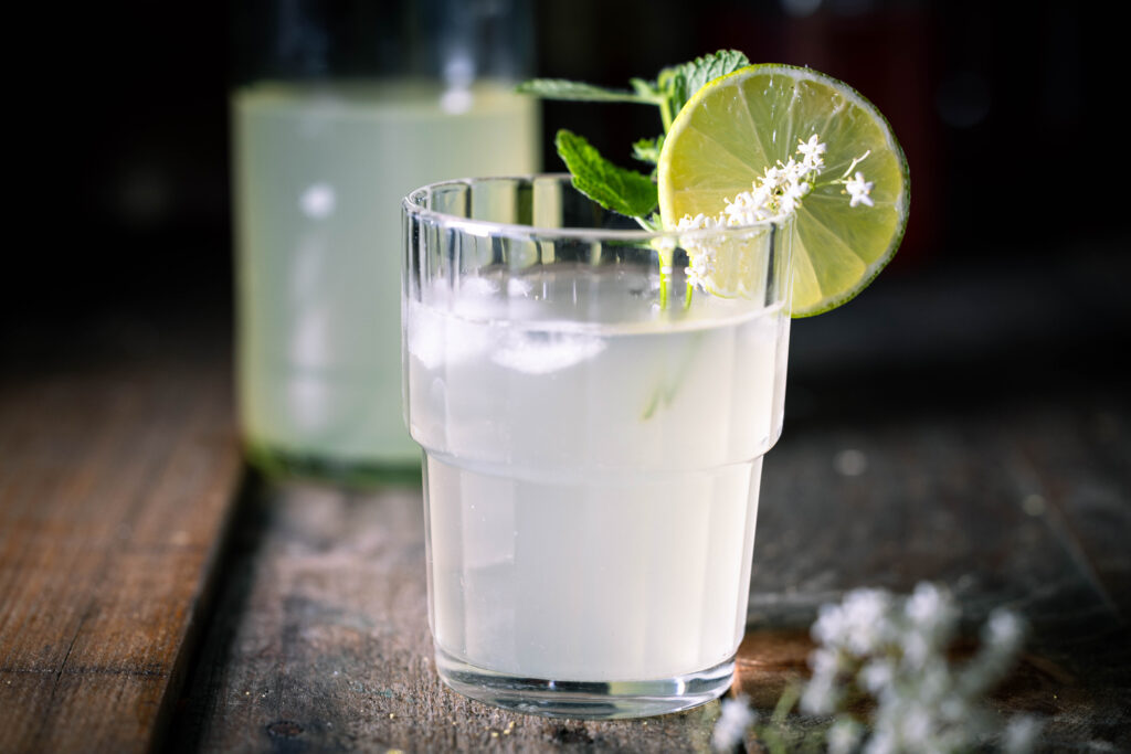 Holunderkefir im Glas mit Eiswürfeln, dahinter in einer angebrochenen Flasche.