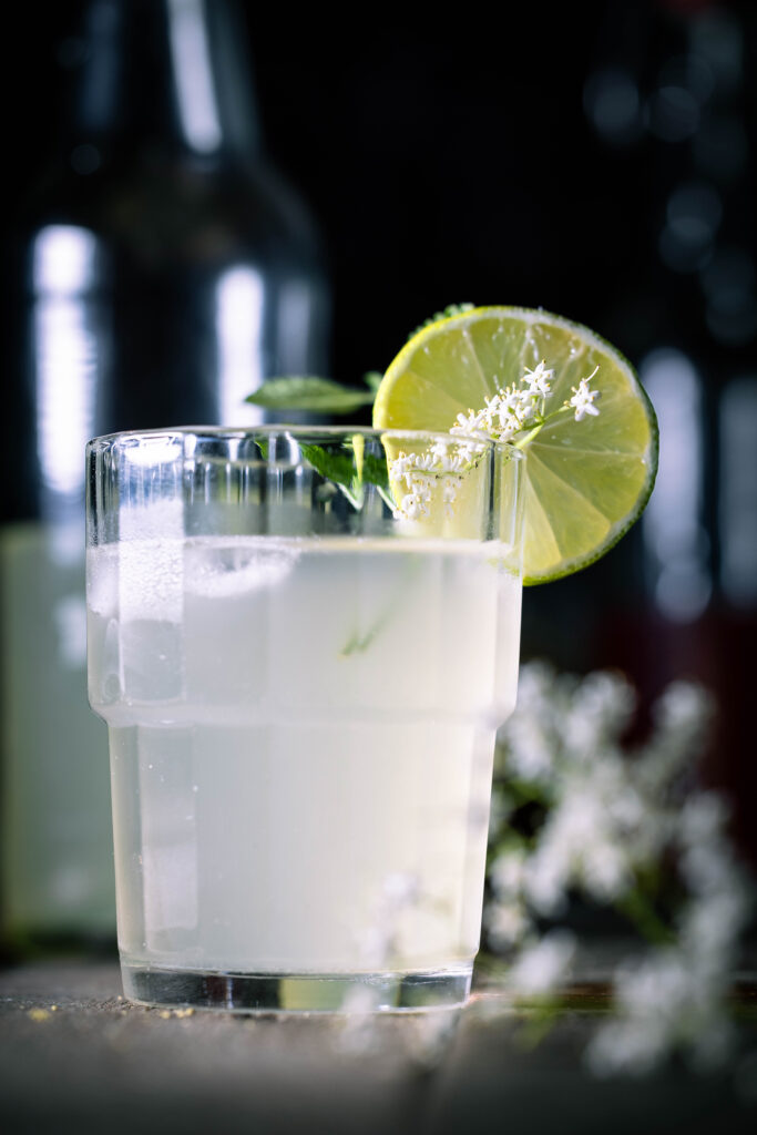 Holunderkefir im Glas mit Eiswürfeln, dahinter in einer angebrochenen Flasche.