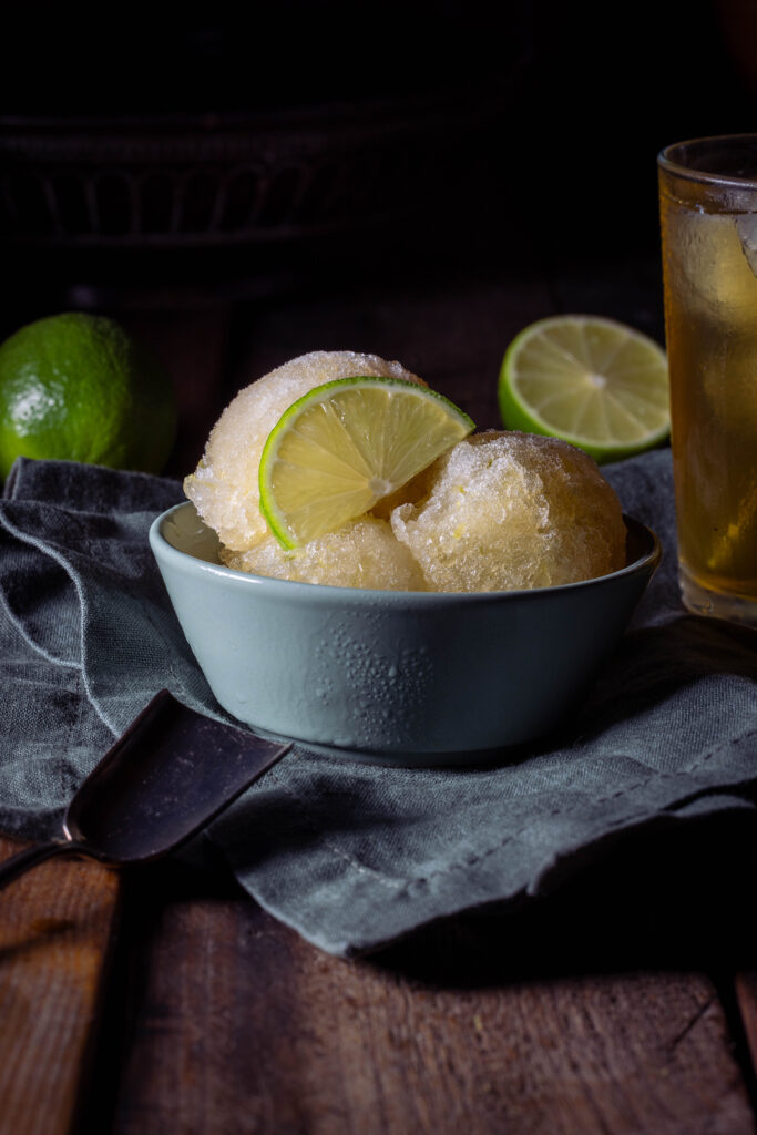 Kombucha-Limetten-Sorbet - drei Kugeln in einer grünen Schale. Dahinter ein Glas eiskalter Kombucha und frische Limetten.