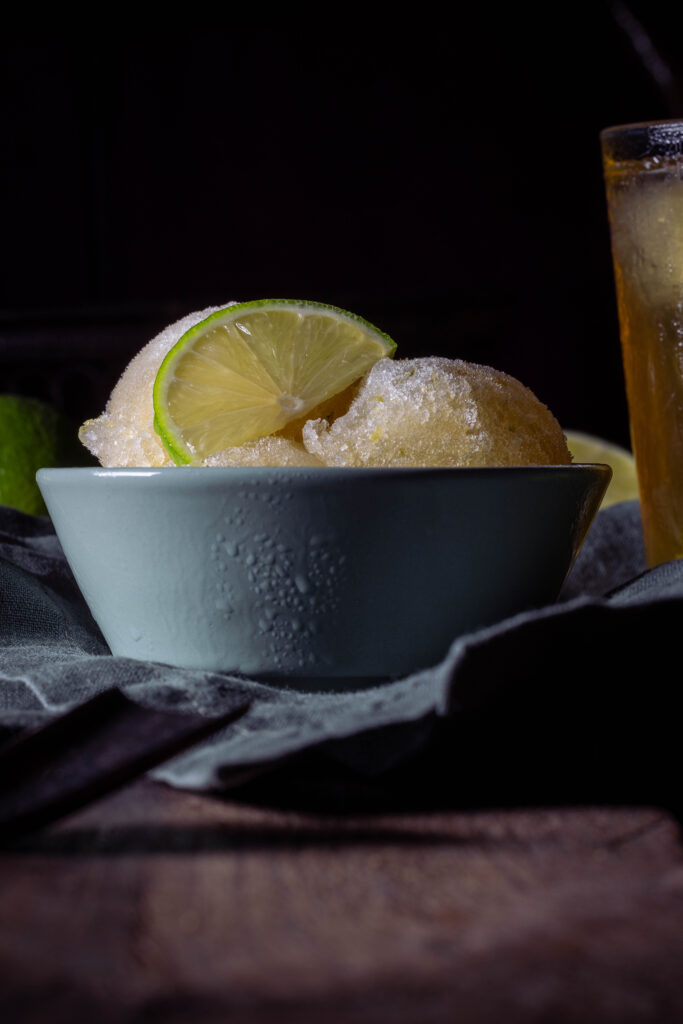 Kombucha-Limetten-Sorbet - drei Kugeln in einer grünen Schale. Dahinter ein Glas eiskalter Kombucha und frische Limetten.