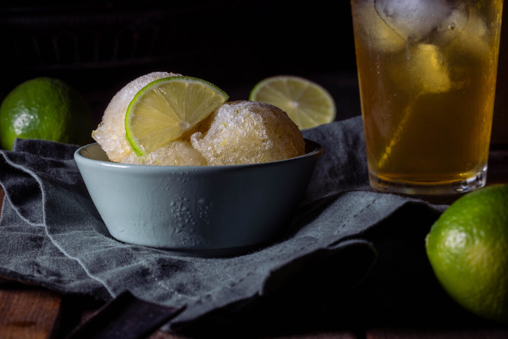 Kombucha-Limetten-Sorbet - drei Kugeln in einer grünen Schale. Dahinter ein Glas eiskalter Kombucha und frische Limetten.
