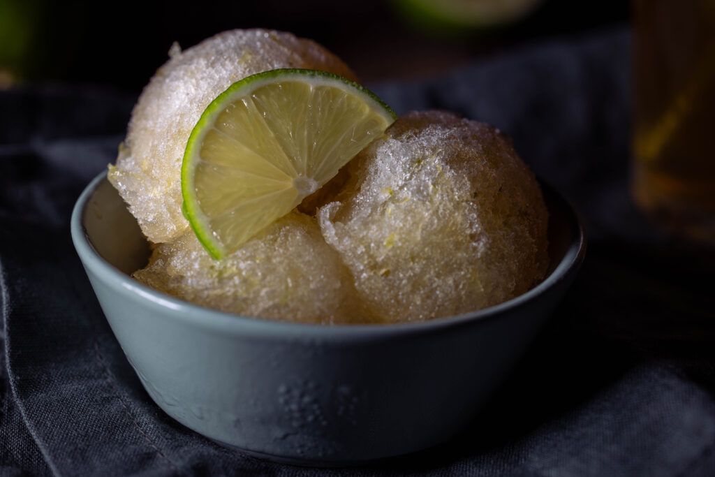 Kombucha-Limetten-Sorbet - drei Kugeln in einer grünen Schale. Dahinter ein Glas eiskalter Kombucha und frische Limetten.