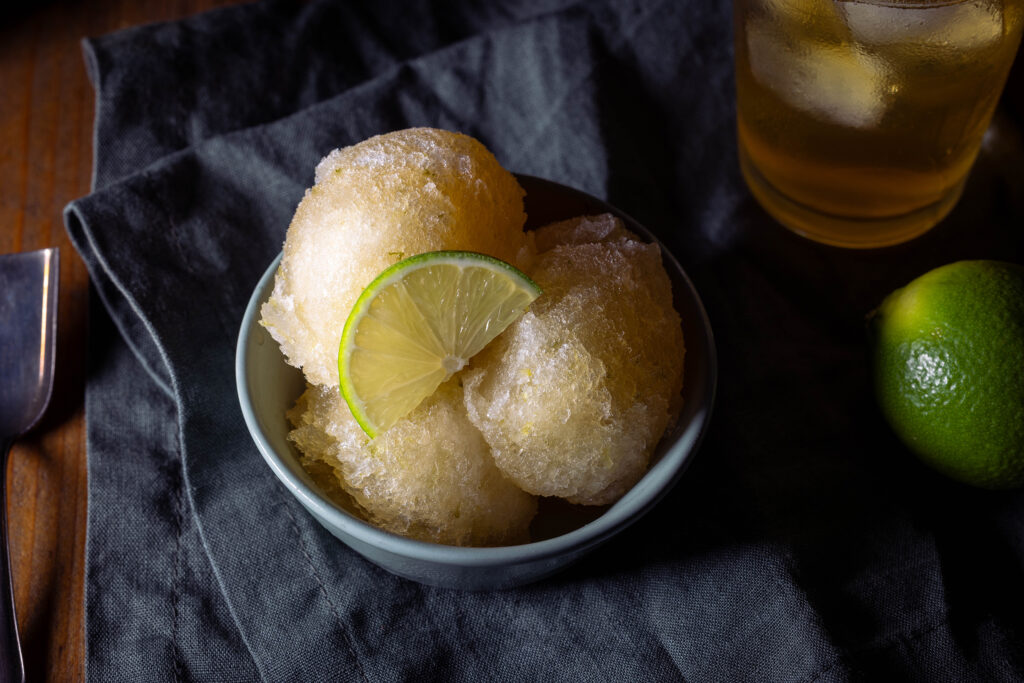Kombucha-Limetten-Sorbet - drei Kugeln in einer grünen Schale. Dahinter ein Glas eiskalter Kombucha und frische Limetten.