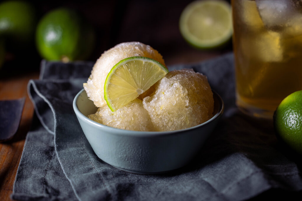 Kombucha-Limetten-Sorbet - drei Kugeln in einer grünen Schale. Dahinter ein Glas eiskalter Kombucha und frische Limetten.