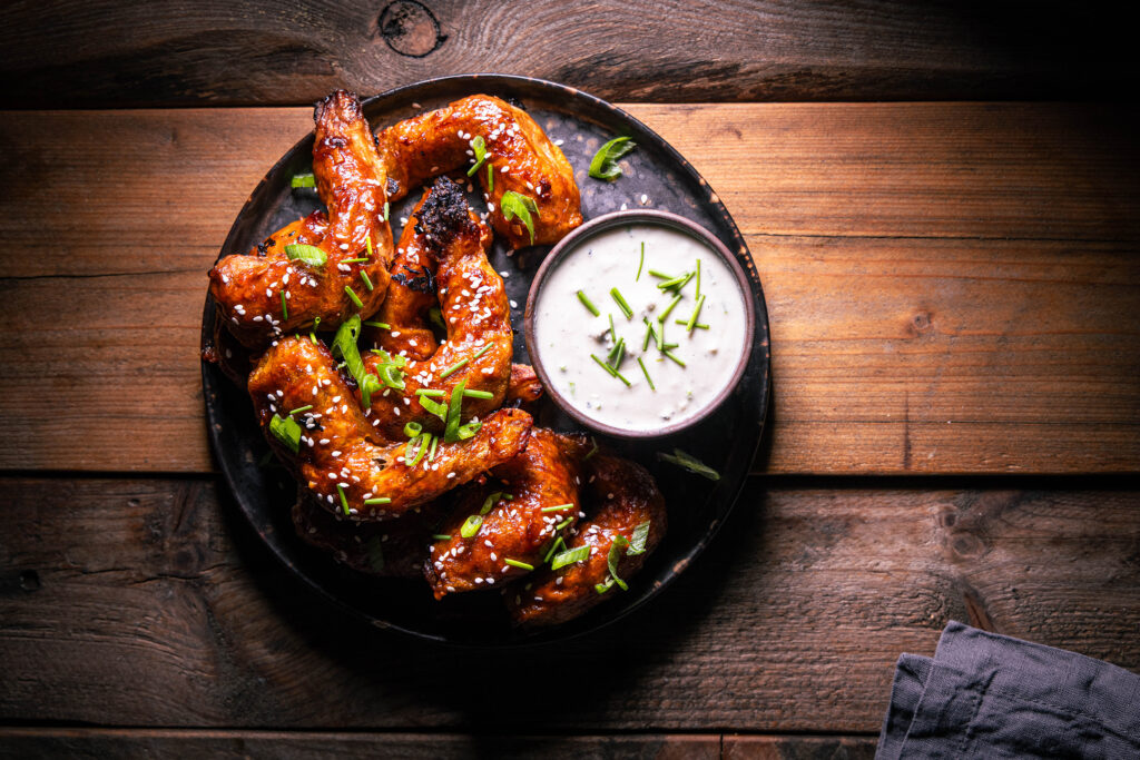 Vegane Sticky Wings - mit Sesam und Lauchzwiebel bestreut auf einem Teller. Außerdem ein Schälchen veganer Baluschimmel-Dip.