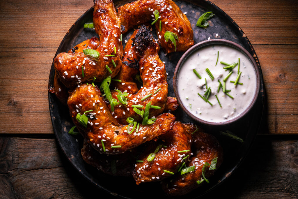 Vegane Sticky Wings - mit Sesam und Lauchzwiebel bestreut auf einem Teller. Außerdem ein Schälchen veganer Blauschimmel-Dip.