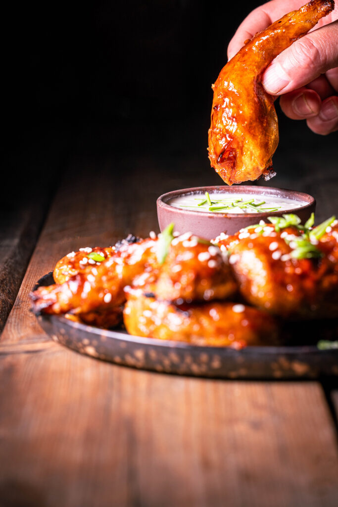 Vegane Sticky Wings – pflanzliche Chicken Wings – Einer wird in einer Hand gehalten, weitere mit Dip auf einem Teller.