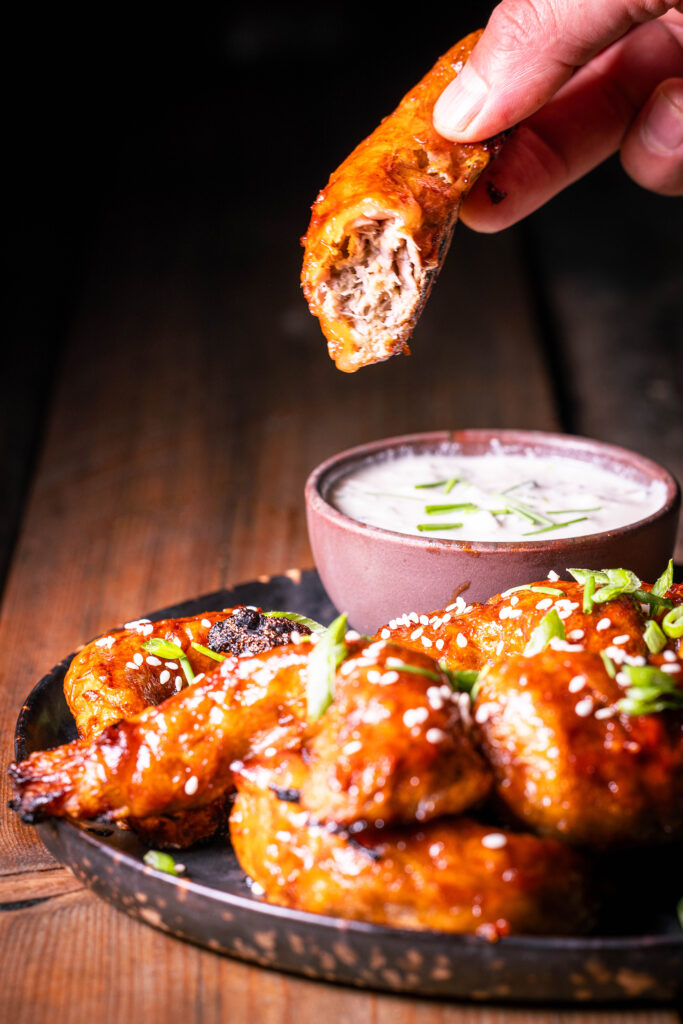 Vegane Sticky Wings – pflanzliche Chicken Wings – Ein angebissener wird in einer Hand gehalten, weitere mit Dip auf einem Teller.