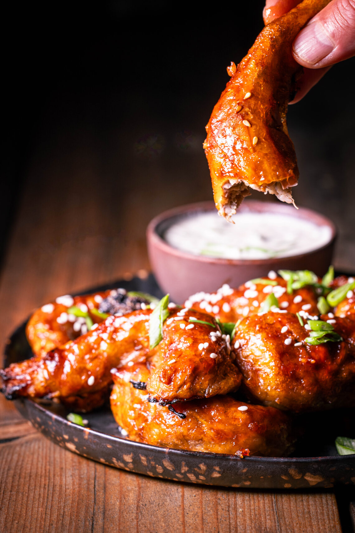 Vegane Sticky Wings – pflanzliche Chicken Wings – Ein angebissener wird in einer Hand gehalten, weitere mit Dip auf einem Teller.