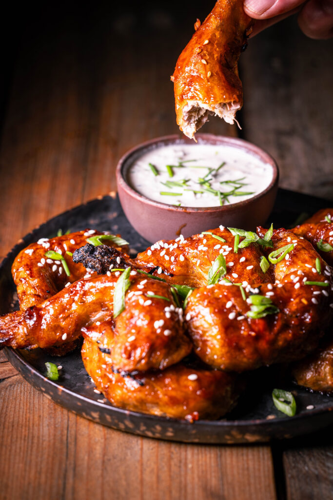 Vegane Sticky Wings – pflanzliche Chicken Wings – Ein angebissener wird in einer Hand gehalten, weitere mit Dip auf einem Teller.