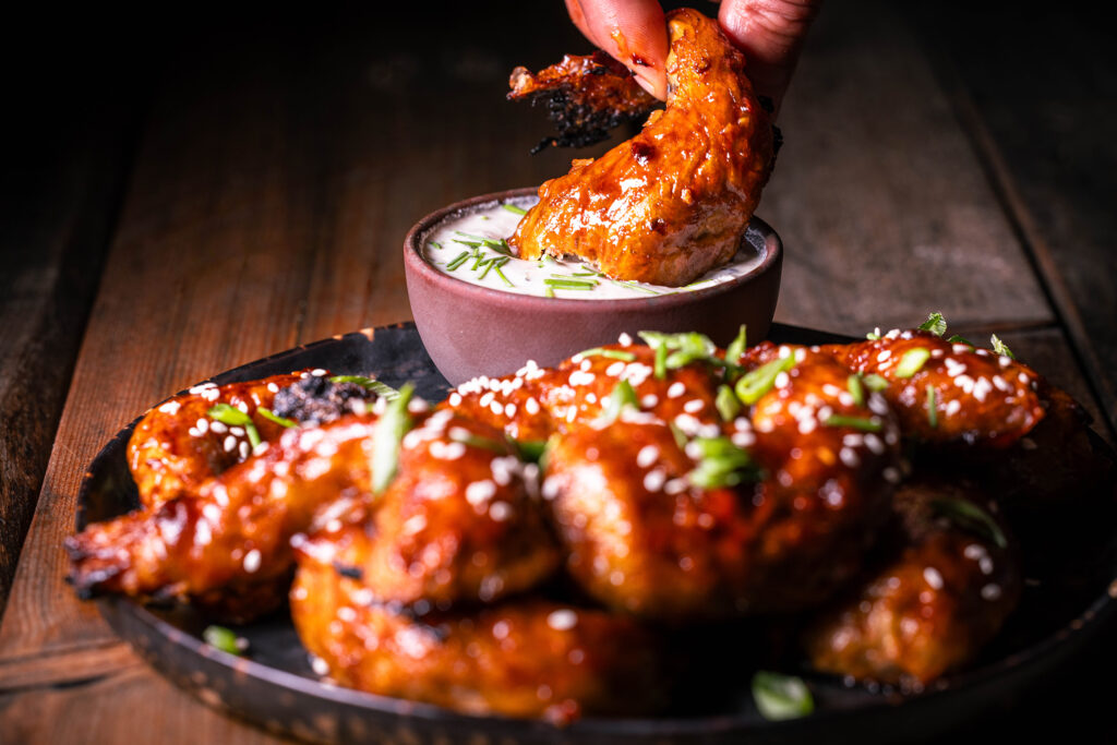 Vegane Sticky Wings – pflanzliche Chicken Wings – Ein angebissener wird in einen Dip getaucht, weitere Wings auf einem Teller.