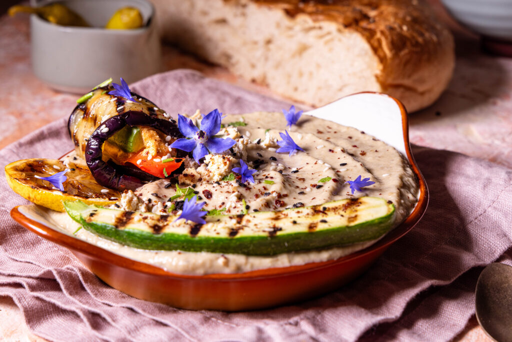 Baba Ganoush in einer Schale, mit Gewürzen, essbaren Blüten, sowie gegrillter Zitrone und Aubergine.