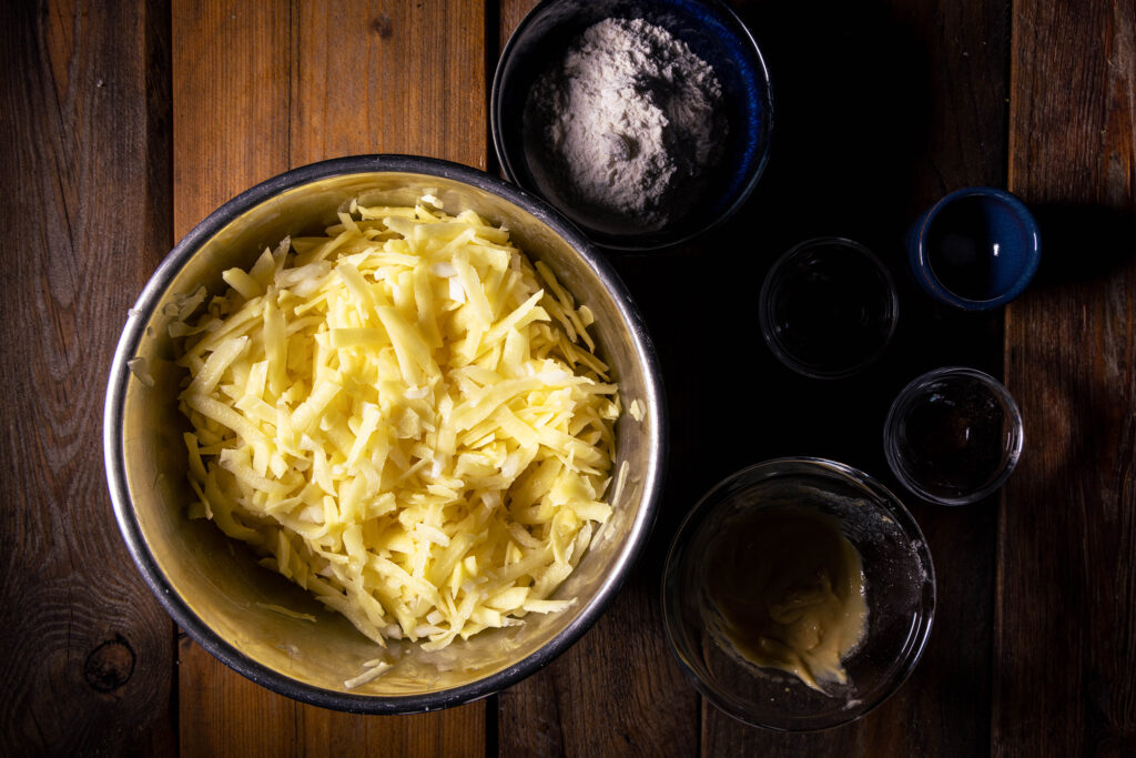 Ein Schüssel mit geriebenen Kartoffeln und gewürfelten Zwiebeln, daneben weitere Zutaten für Kartoffelpuffer.