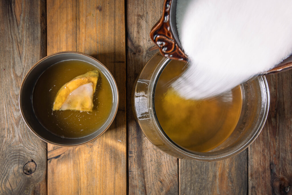 Zucker wird in ein Glasgefäß mit Mate-Tee gekippt. Daneben eine Schale mit einem Scoby.