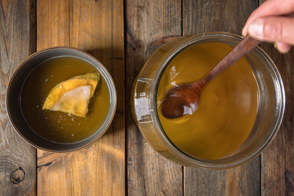 Zucker wird in ein Glasgefäß mit Mate-Tee gerührt. Daneben eine Schale mit einem Scoby.