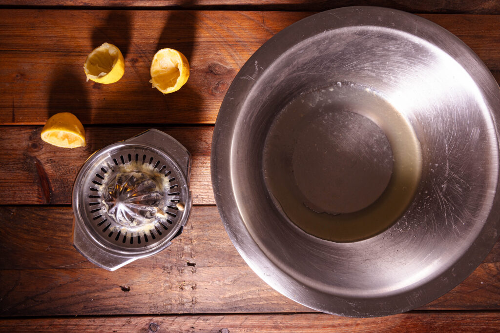 Eine Zitronenpresse, drei ausgepresste Zitronenhälften und eine Schüssel mit Zitronensaft darin.